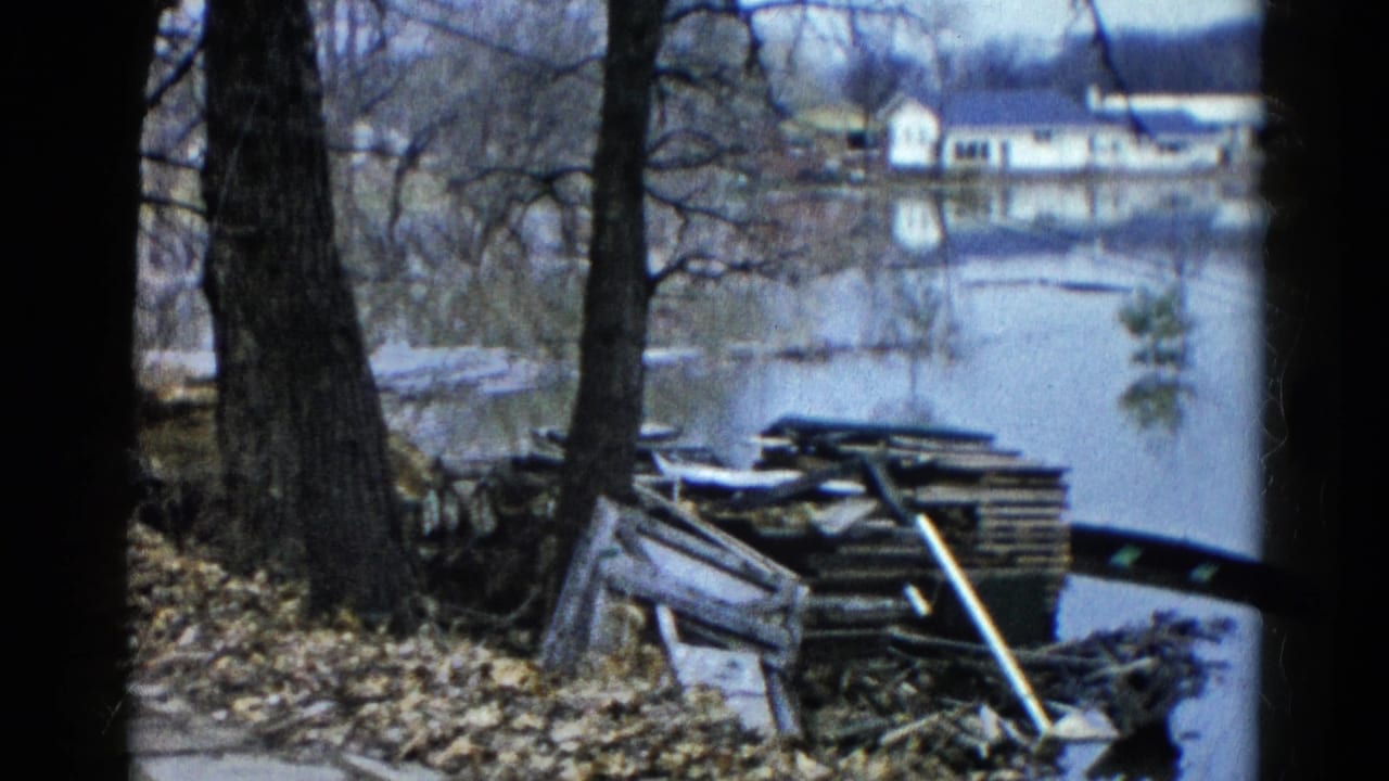 1961 Wauconda 8mm frame showing floodwater near bare trees, shoreline debris, stacked wood, and houses beyond the water.