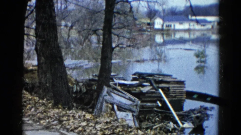1961 Wauconda 8mm frame showing floodwater near bare trees, shoreline debris, stacked wood, and houses beyond the water.