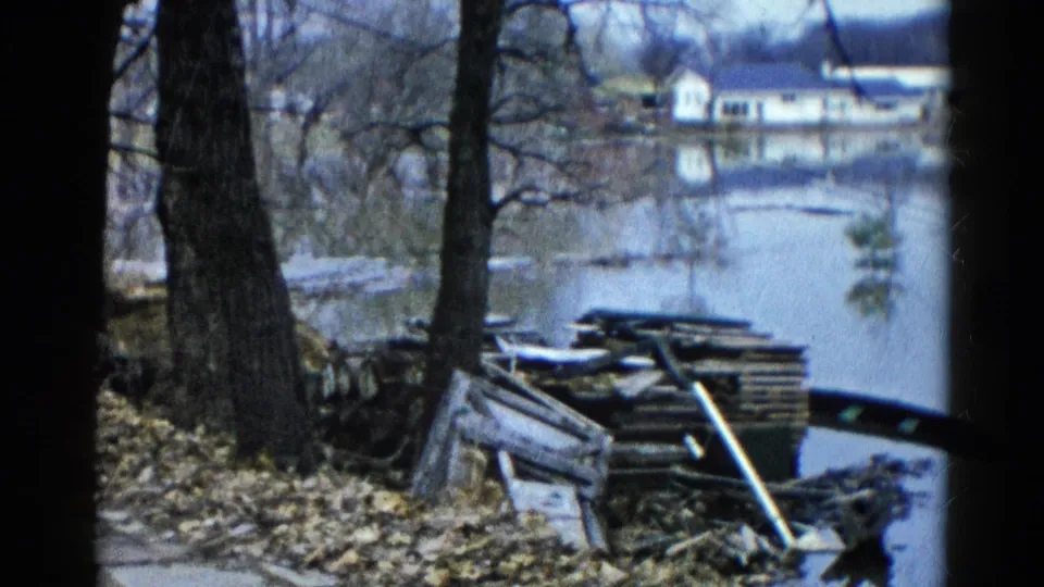 1961 Wauconda home-movie frame of a flooded shoreline with bare trees, stacked lumber, scattered debris, and distant houses.