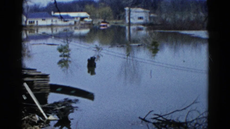 1961 Wauconda 8mm frame showing broad floodwater, distant houses, poles, tree reflections, branches, and debris at the lower edge.