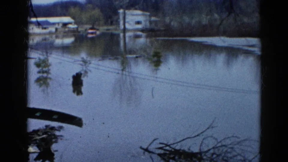 1961 Wauconda home-movie frame showing floodwater across a residential area, with distant houses, poles, bare branches, and debris.