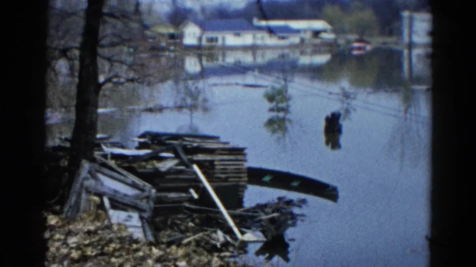 1961 Wauconda, Illinois flood view with a pile of planks in the foreground and residential buildings reflected across the water.
