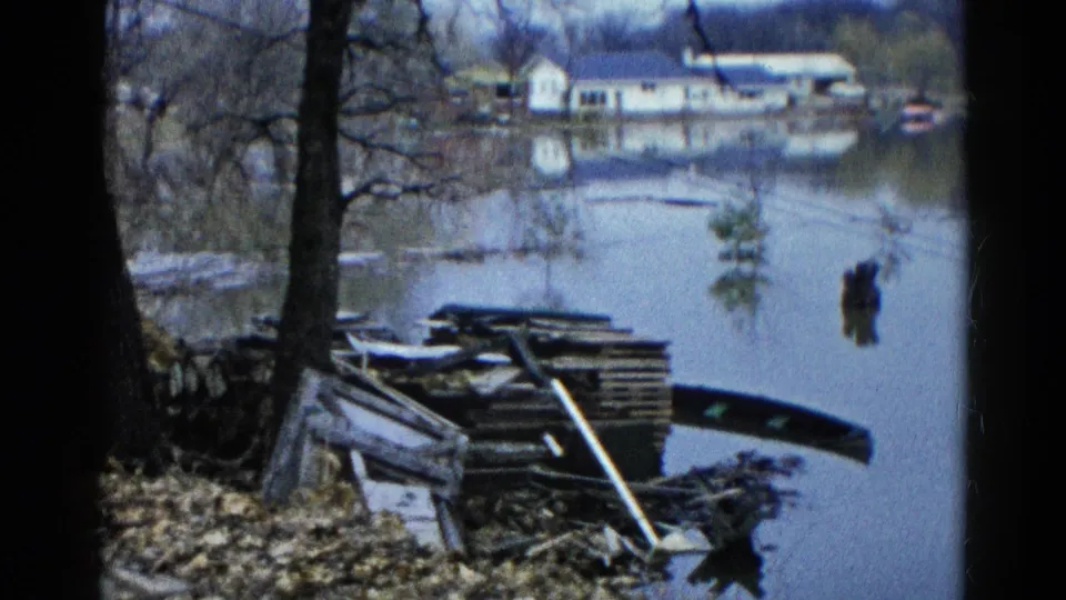 1961 Wauconda, Illinois 8mm frame showing bare shoreline trees, a debris pile, calm floodwater, and houses in the distance.