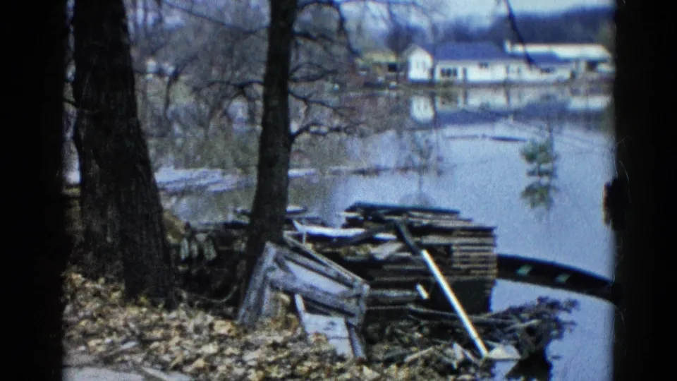1961 Wauconda, Illinois flooded neighborhood view with foreground tree trunks, stacked debris, and reflected buildings across still water.