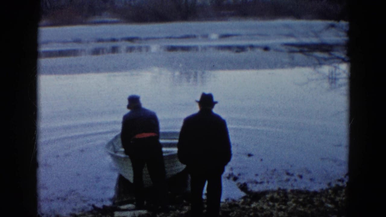 Two men stand beside a small rowboat at the edge of a calm lake.