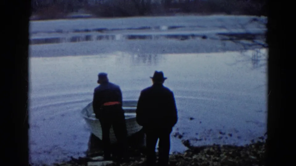 Two adult men in dark coats stand near a rowboat on a calm lakeshore in Wauconda, Illinois, in 1961 home-movie footage.