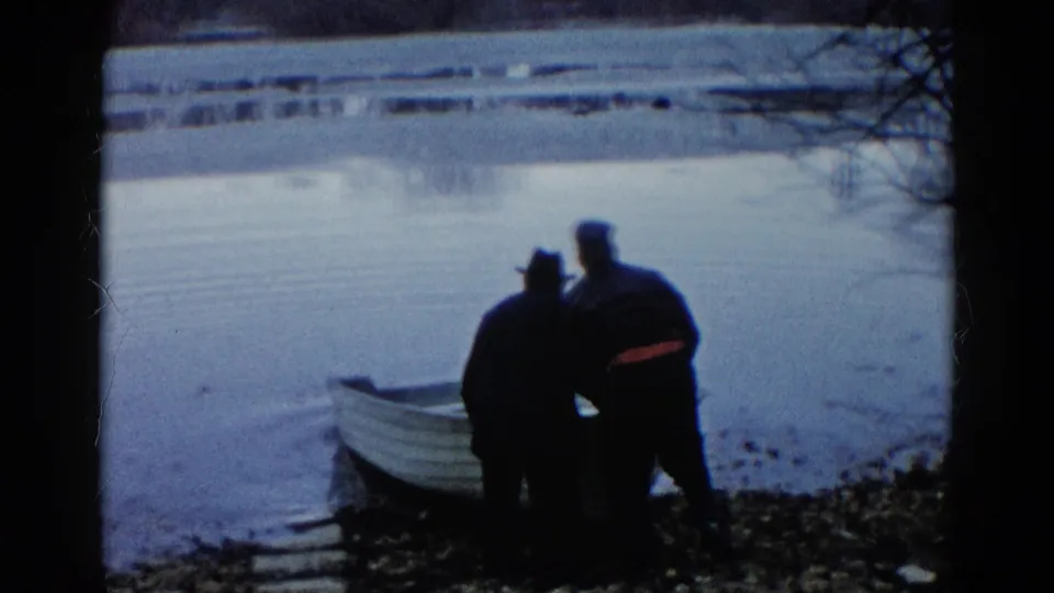 In Wauconda, Illinois, two men in dark coats and hats stand near a white rowboat at the edge of a calm lake.