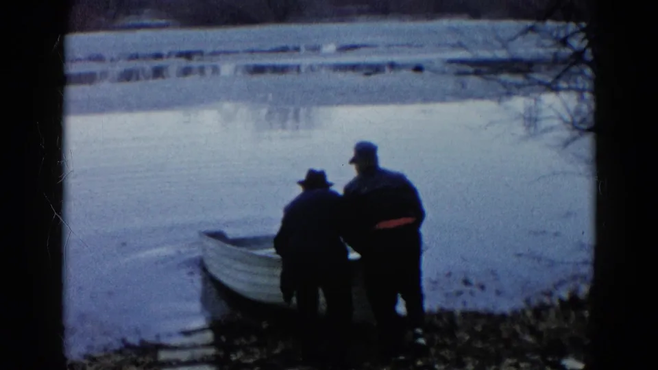 Two silhouetted men in dark outerwear stand beside a small rowboat on a quiet Wauconda lakeshore.