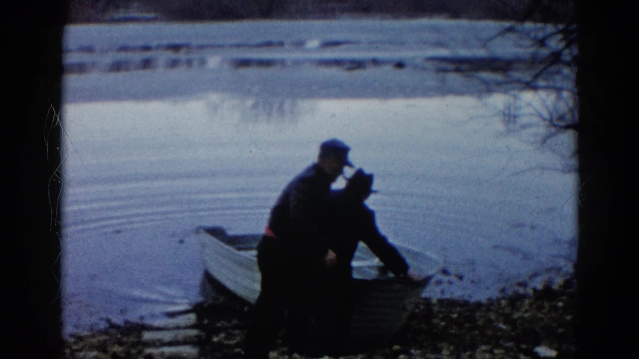 In Wauconda, Illinois, 1961, two adults in dark jackets and hats stand by a small rowboat at the edge of a calm lake.