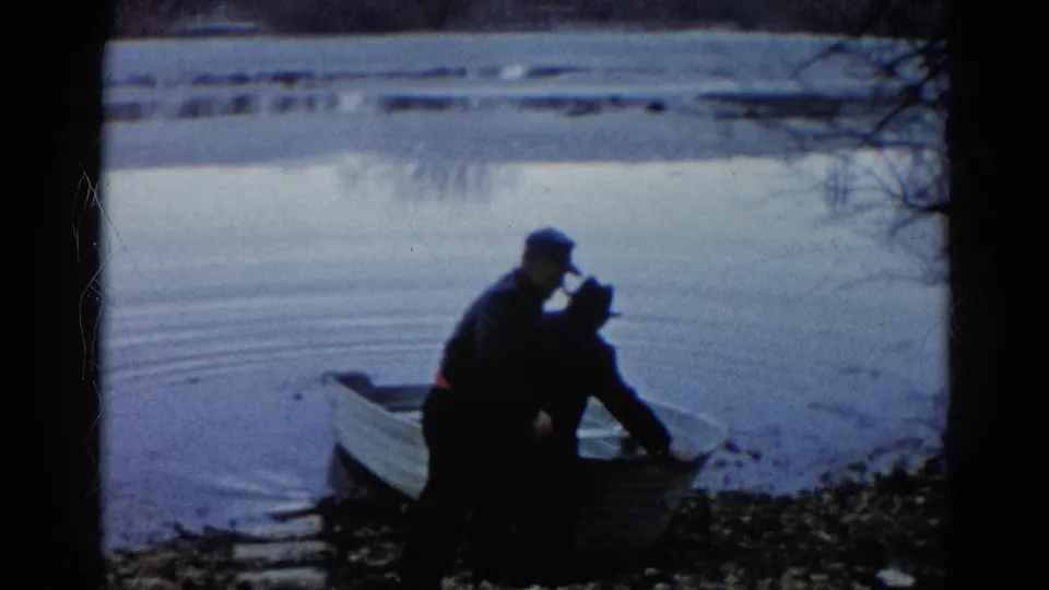 In Wauconda, Illinois, 1961, two adults in dark jackets and hats stand by a small rowboat at the edge of a calm lake.