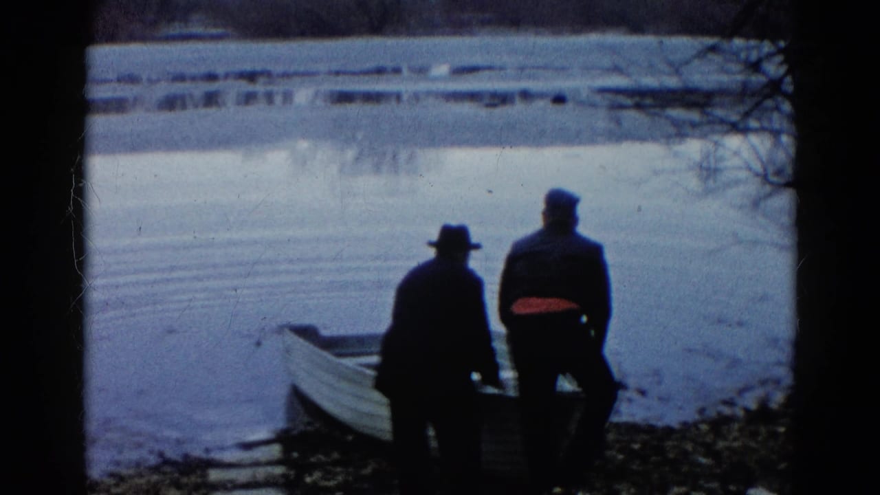 Two adults in hats and jackets stand close to the bow of a small rowboat on the Wauconda lakeshore in 1961.
