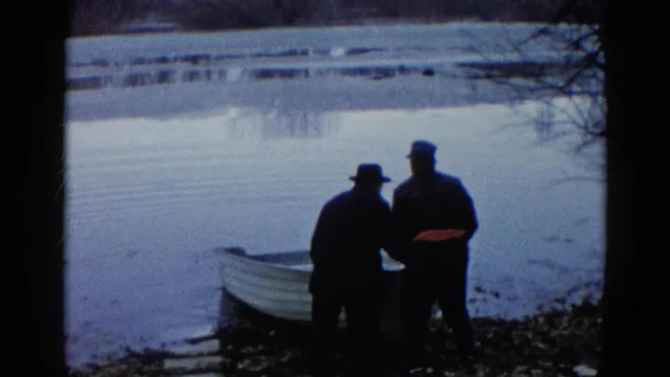 Two adults remain beside the rowboat at the calm Wauconda lakeshore, framed by dark branches and 8mm film edges.