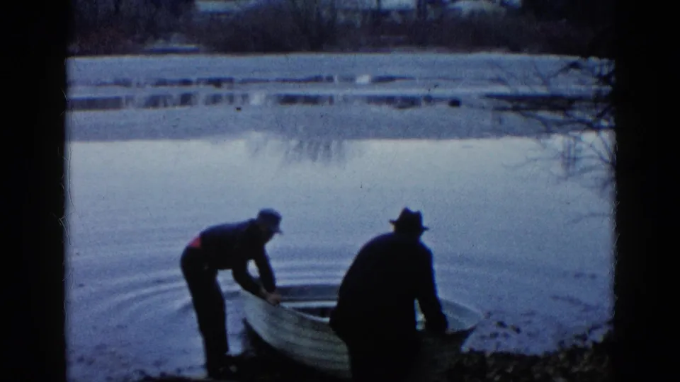 Two adults in dark clothing work beside a small rowboat at the Wauconda lake edge, with ripples visible on the water.