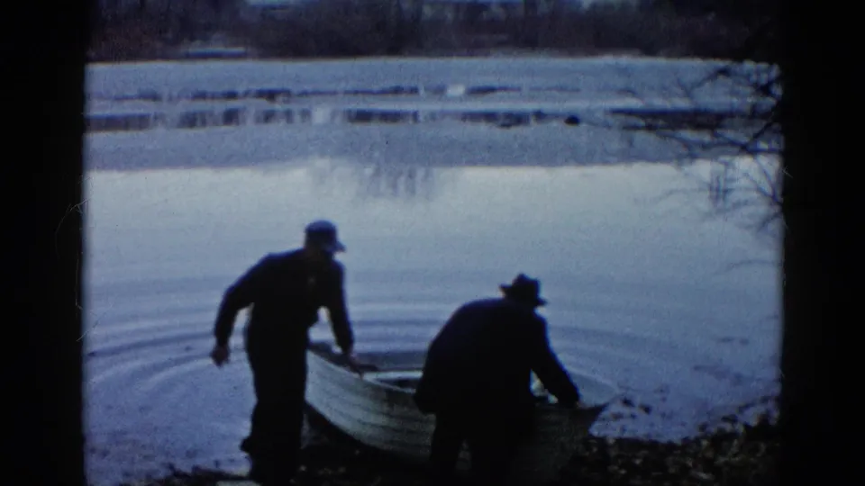 At the Wauconda lakeshore, one capped man stands near the stern while another hatted man bends over the rowboat at the waterline.