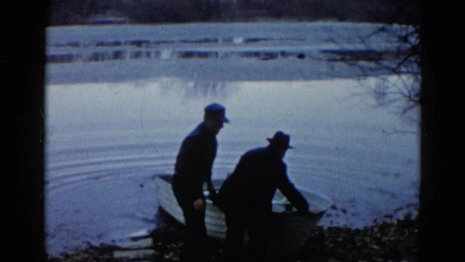 Two silhouetted men remain beside the small rowboat at the Wauconda, Illinois lake edge, with smooth water and a distant shore behind them.