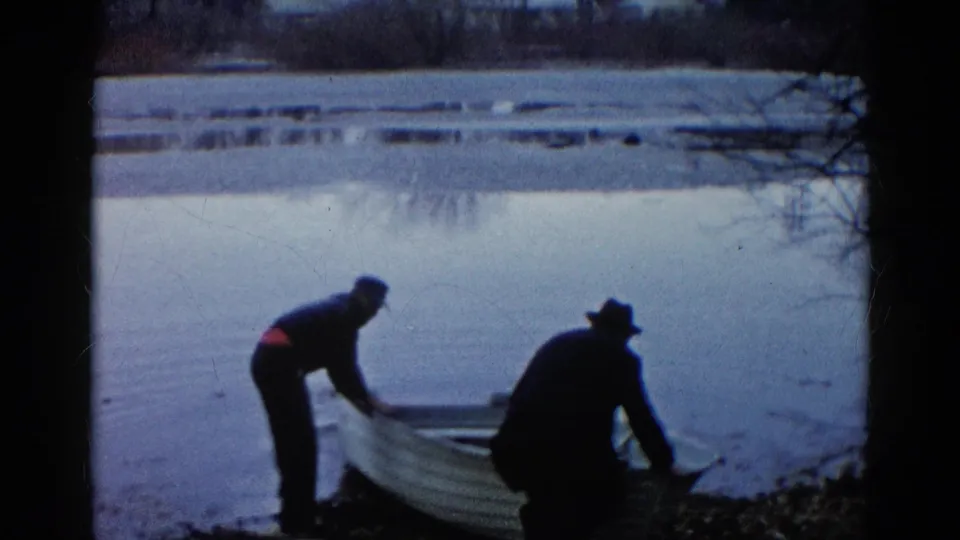 Two men stand on the shore as the rowboat extends into the calm lake, framed by dark trees.
