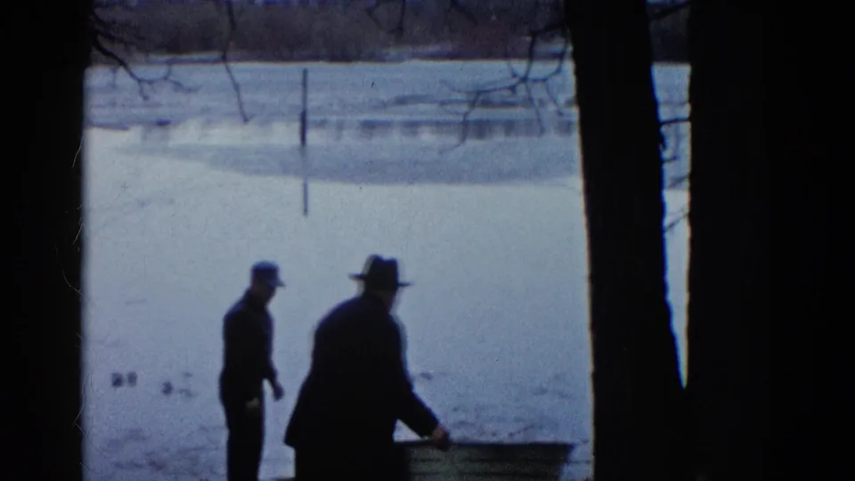 Two men in hats stand near a rowboat on a snow-covered Wauconda lakeshore, with the frozen lake behind them.