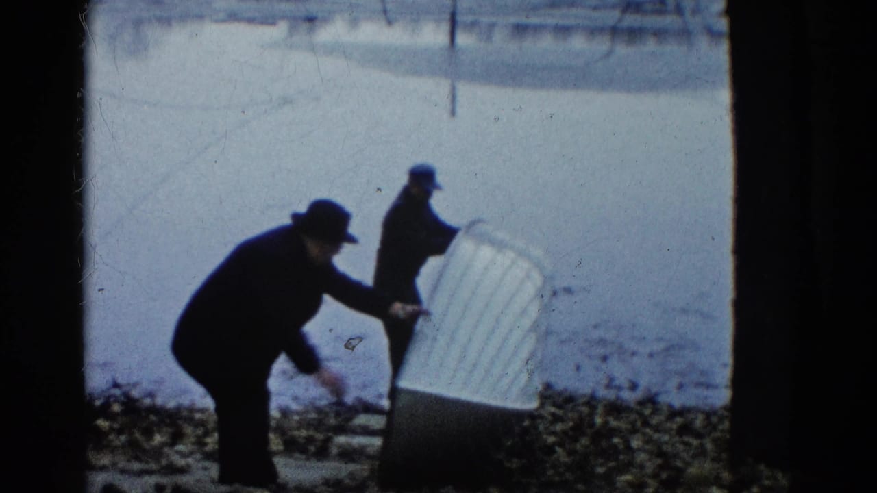 Two men in dark coats handle a small white rowboat on a pebbled shore beside calm water.