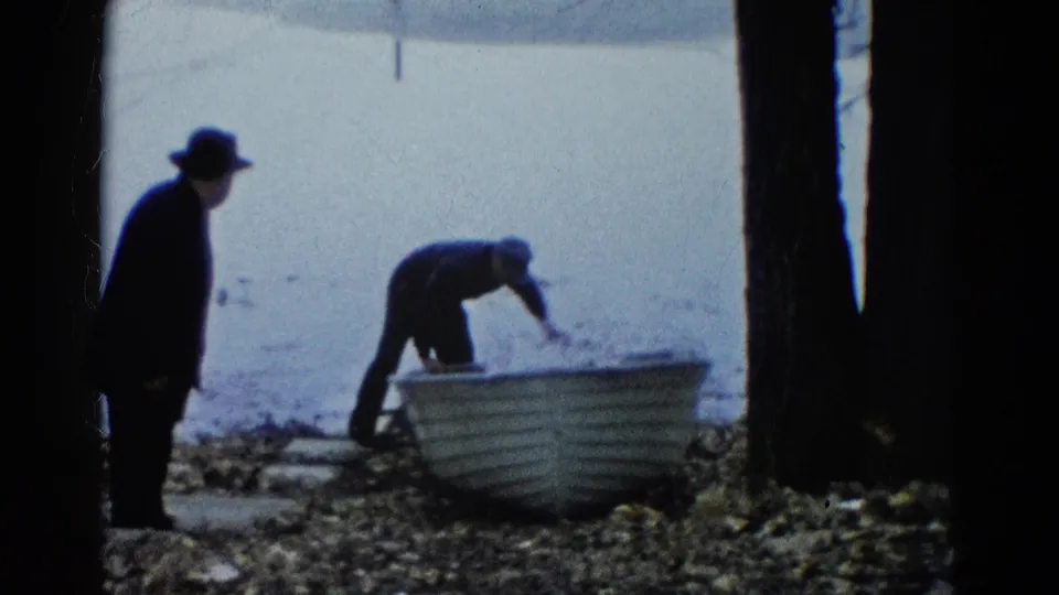 A 1961 Wauconda lakeside frame shows one man watching as another adjusts a small white rowboat near the waterline.