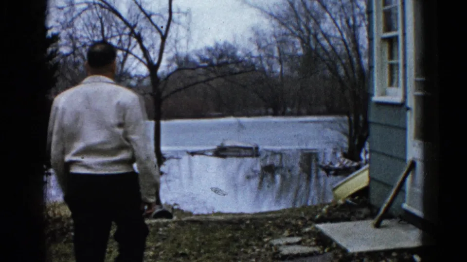 In Wauconda, Illinois, 1961, a man in a light sweater walks from a house toward floodwater beyond the yard.