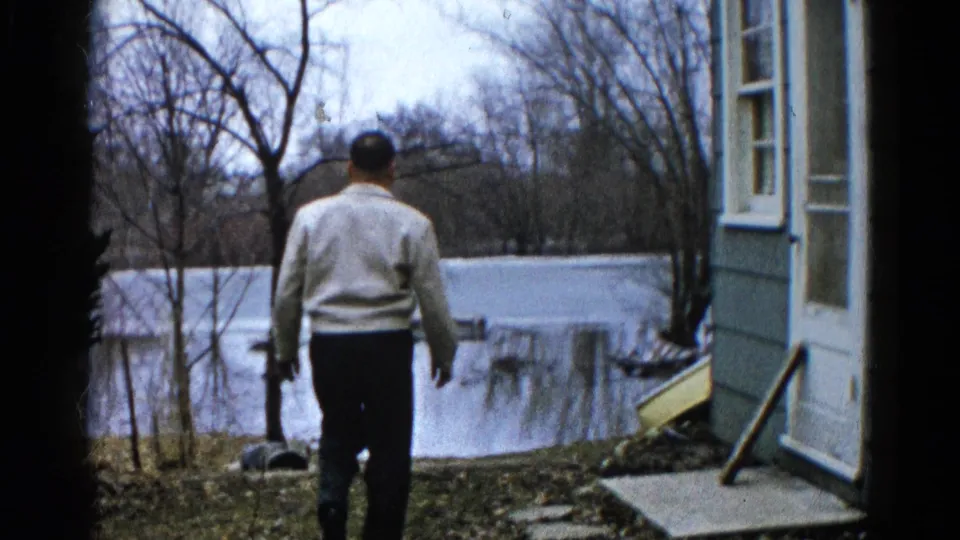 The man continues toward the water's edge beside a house, with bare trees and a flooded field visible in Wauconda, Illinois.