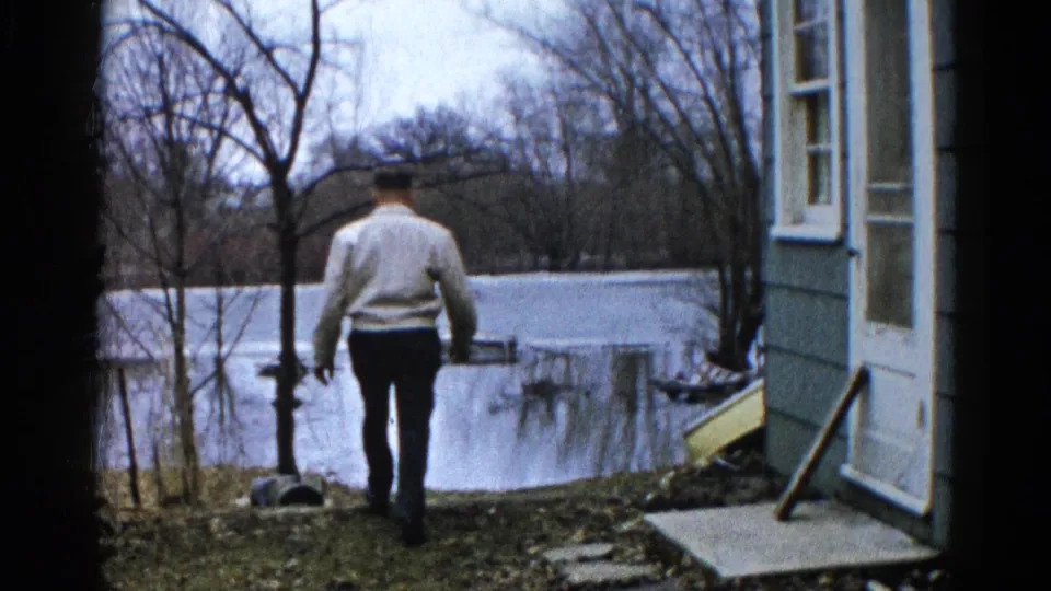 In the 1961 Wauconda home movie, the man reaches the edge of a flooded yard near a house and leafless trees.