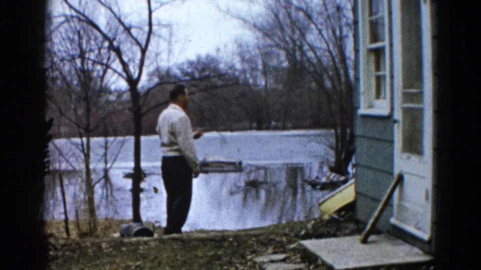 The man pauses near the floodwater in profile, with the house wall, bare trees, and partially submerged yard behind him.