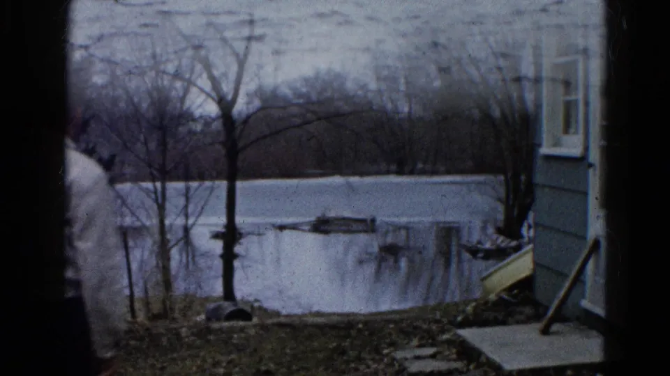 1961 Wauconda frame showing an older man in a light sweater near a winter lakeside yard, with a house and small boat by icy water.