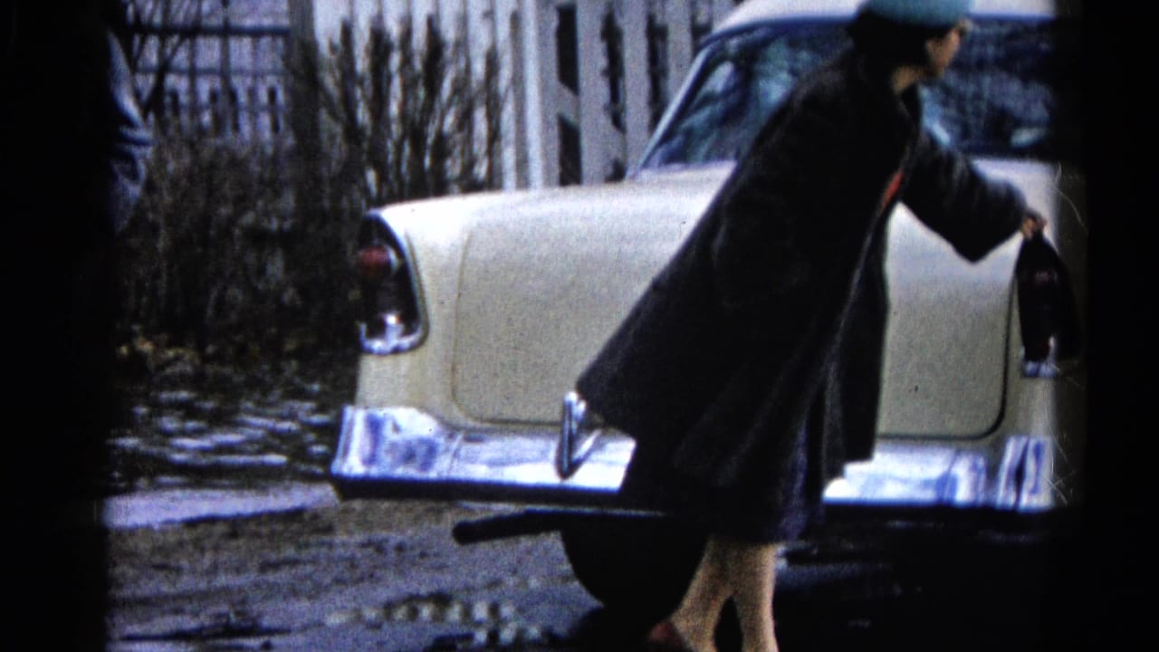 In 1961 Wauconda, Illinois, a woman in a dark coat and hat stands near the rear fender of a cream classic car on a muddy, wet street.