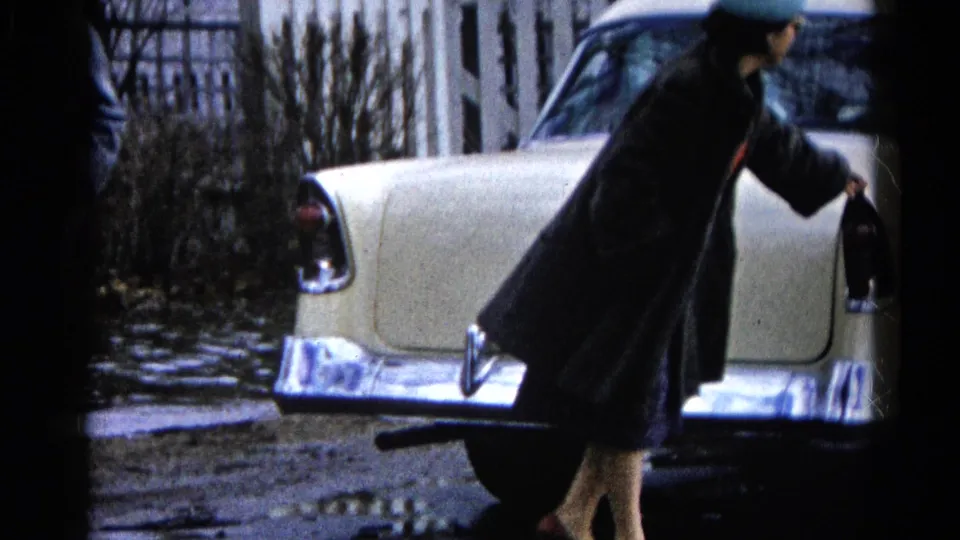 In 1961 Wauconda, Illinois, a woman in a dark coat and hat stands near the rear fender of a cream classic car on a muddy, wet street.