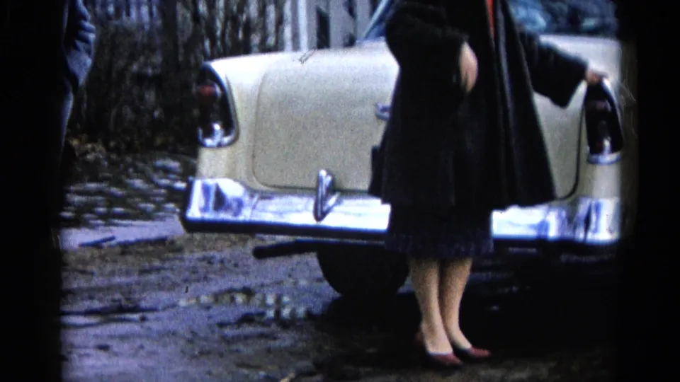 A woman in a dark coat and red heels stands beside a cream classic car on a wet Wauconda residential street, consistent with the 1961 flood setting.