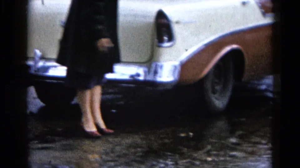 The frame shows the woman’s coat and heels next to the cream-and-tan car, with wet pavement and standing water visible around the rear wheel area.