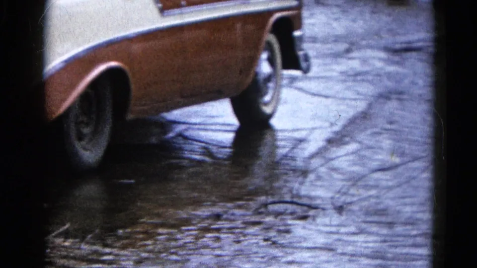 A cream-and-tan classic car fills the frame on a wet residential street, with puddles and reflective floodwater visible along the curb area.