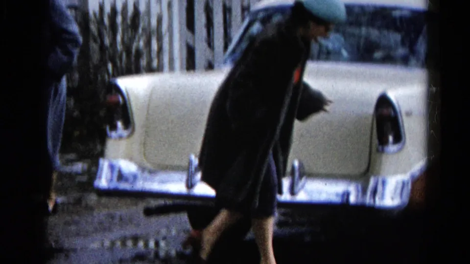 A woman in a dark coat and skirt walks toward the rear of the classic Chevrolet, with red shoes visible against the wet street.