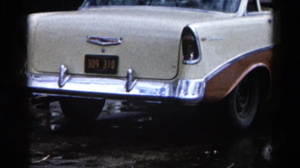 Close 8mm view of the Chevrolet rear, chrome bumper, and Illinois plate beside wet shoreline ground.