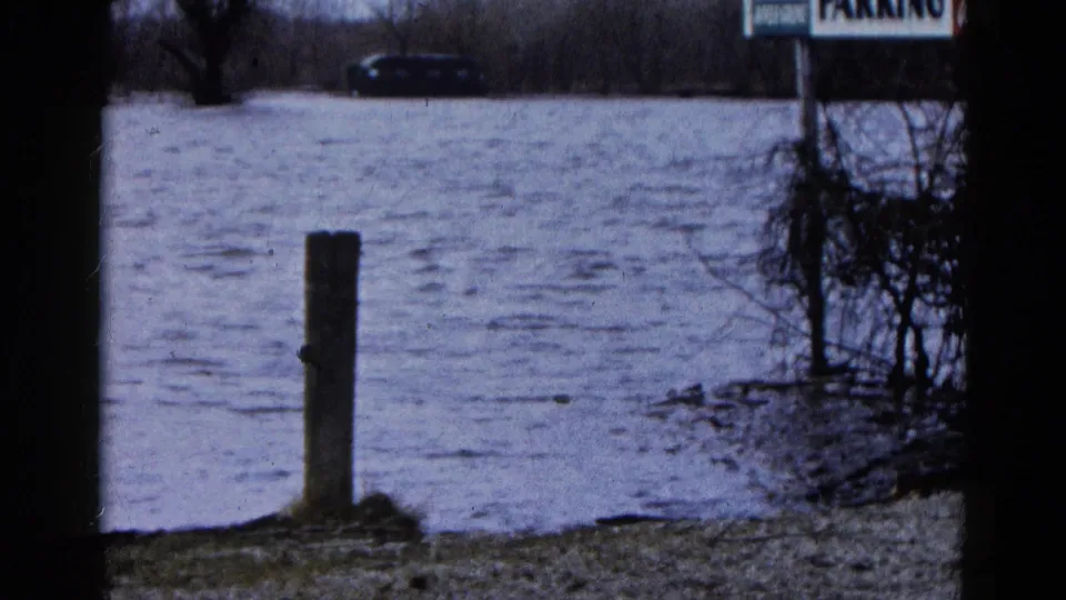 In Wauconda, Illinois, choppy 1961 floodwater reaches a parking area with a foreground post, brush, sign, and partly submerged car.