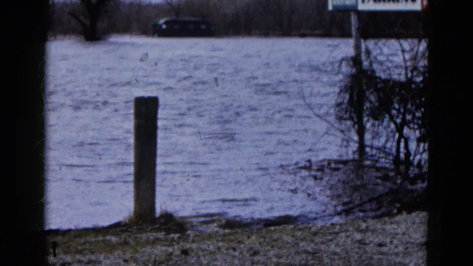 1961 Wauconda flood footage shows water across a parking area, a dark car in the distance, and a foreground post near brush.