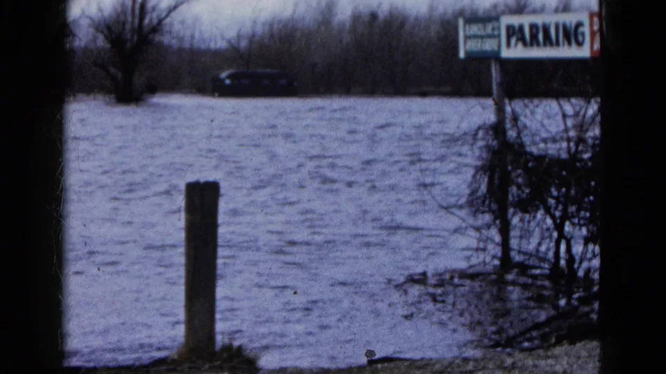 1961 home-movie view of Wauconda floodwater reaching a wooden post and roadside parking sign, with trees and a distant car behind.