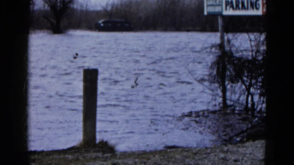 Wauconda, Illinois floodwater in 1961 with a foreground post, bare trees, brush, a distant car, and a cropped parking sign.