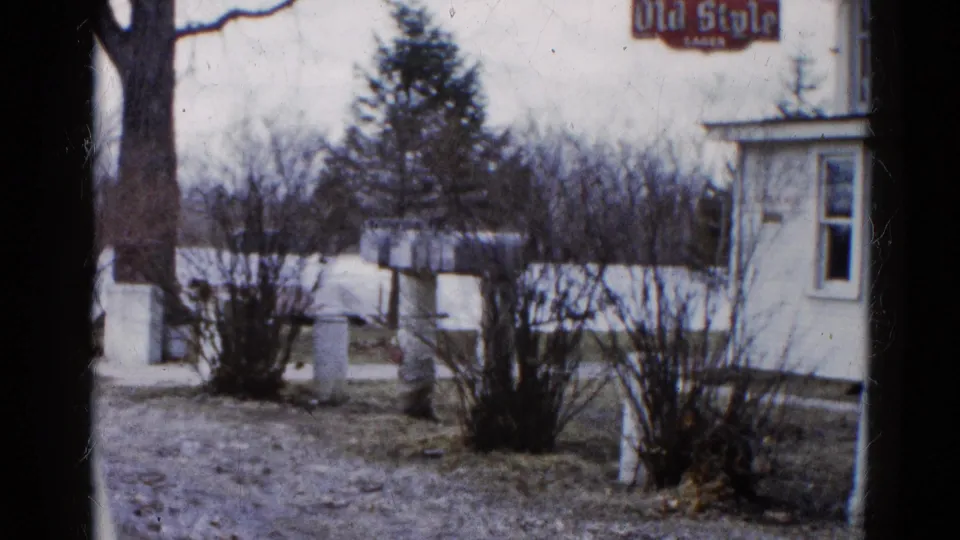 1961 Wauconda, Illinois frame with the edge of a white roadside building, Old Style signage, winter-bare shrubs, and floodwater nearby.