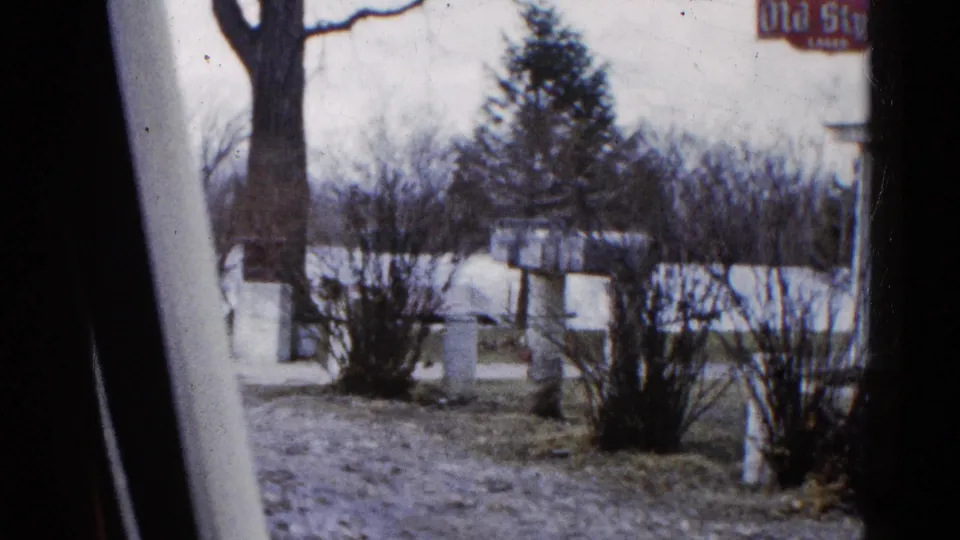 1961 Wauconda, Illinois home-movie frame looking past a tree or vehicle edge toward shrubs, floodwater, and partial Old Style signage.