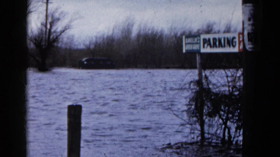 1961 Wauconda, Illinois frame showing a submerged parking area with a PARKING sign, roadside post, brush, and a dark car beyond the water.