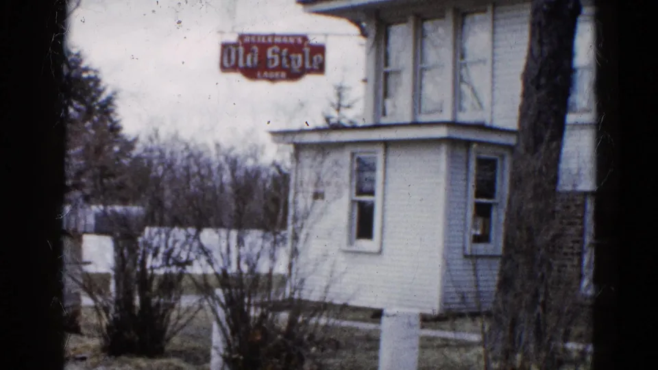 1961 Wauconda, Illinois 8mm frame showing a white house exterior with a red Old Style Lager sign, winter shrubs, and a tree trunk at right.