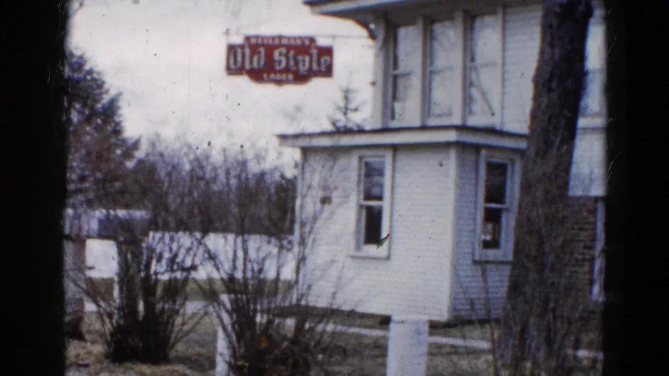 1961 Wauconda, Illinois 8mm still showing the house corner, porch windows, Old Style Lager sign, bare shrubs, and muted winter yard.