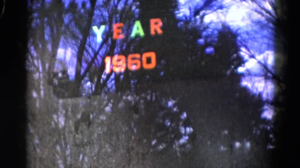 Low-light Wauconda home-movie frame showing colored YEAR 1960 lights partly softened by motion blur and tree silhouettes.