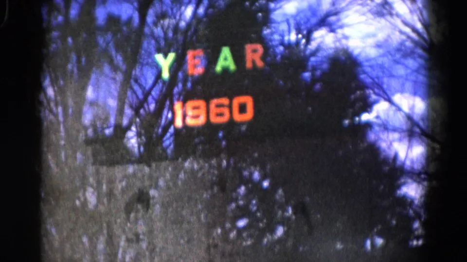 Reflected YEAR 1960 sign in early 1960s Wauconda footage, with branches and dark foliage behind it.