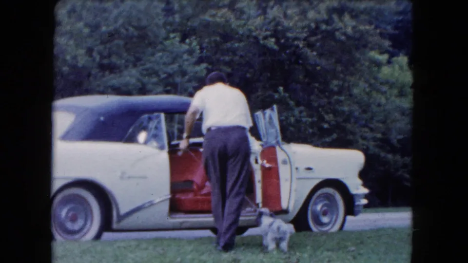In 1961 South Carolina home-movie footage, a man stands by an open white convertible as a small dog waits on the grass.
