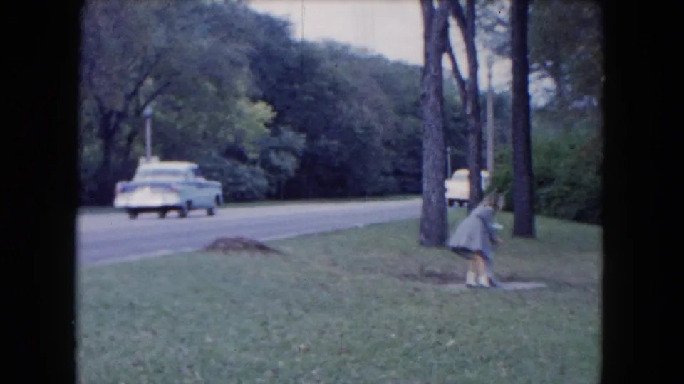 A child near the roadside grass looks toward the ground while a white mid-century car passes on the tree-lined road.