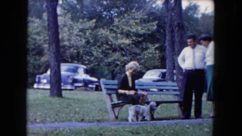 1961 South Carolina park frame with a small dog walking near a green bench, a seated woman, standing adults, trees, and parked cars.