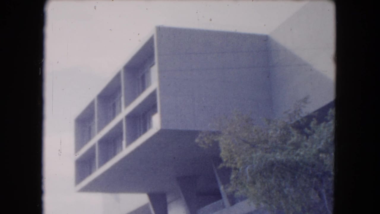 Cantilevered white building corner with recessed windows, trees, and pale sky.